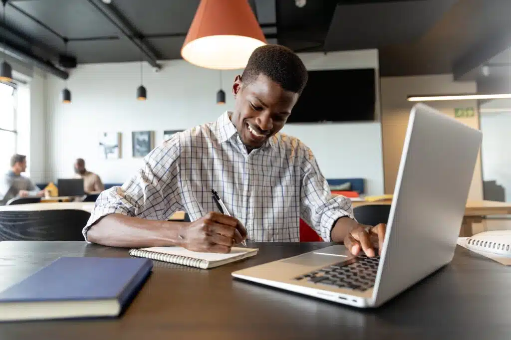Smiling young african american businessman writing in diary and using laptop in creative office