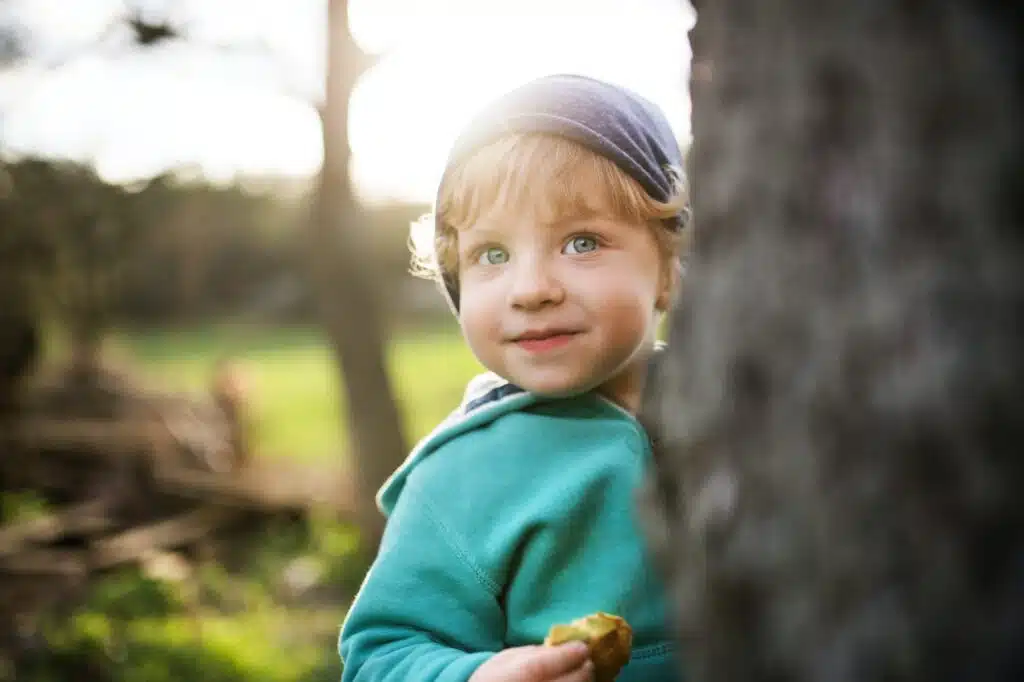 A happy toddler boy hiding behind tree outside in spring nature.