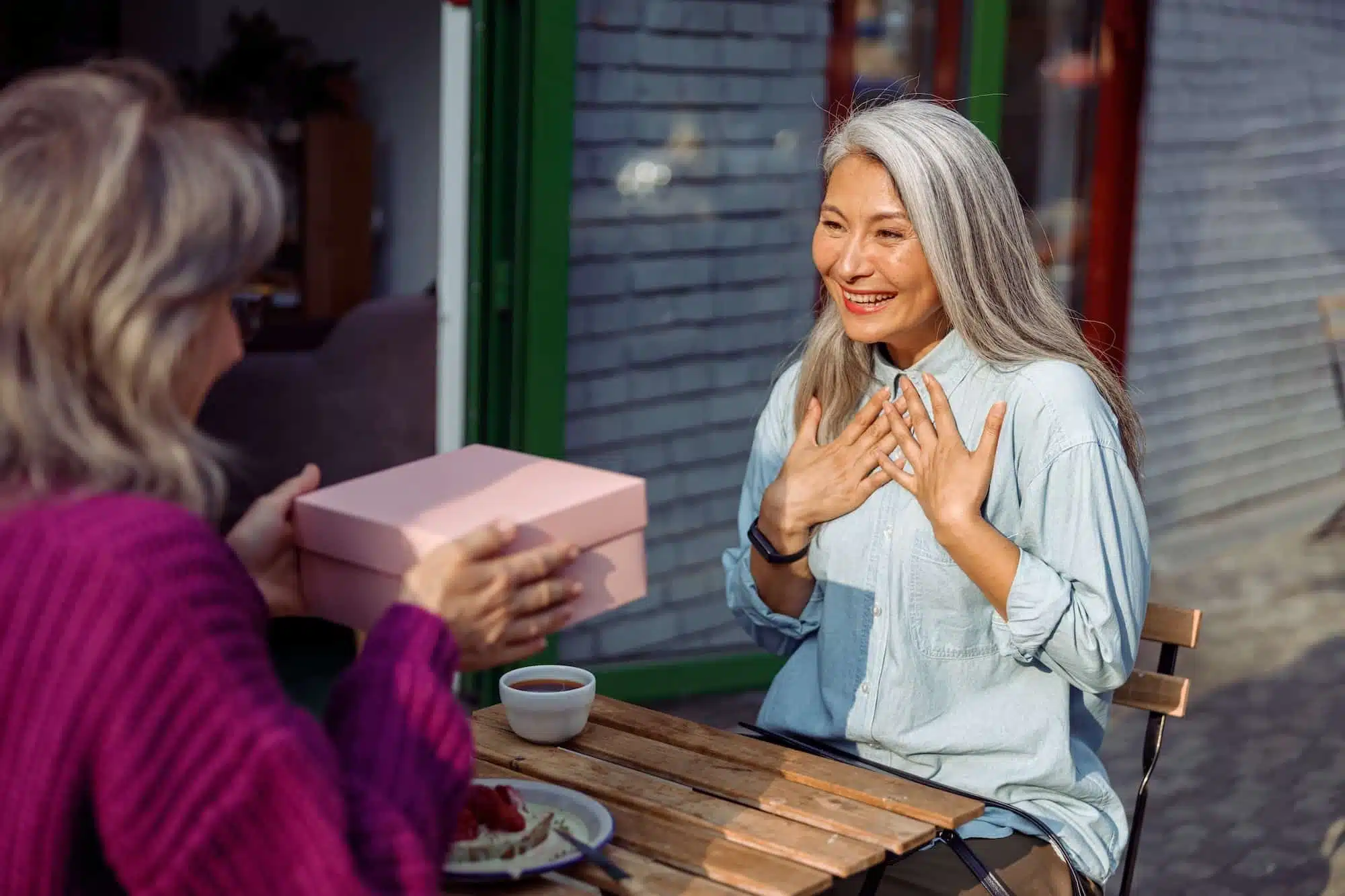Mature friend gives present to emotional Asian woman sitting at small table in cafe