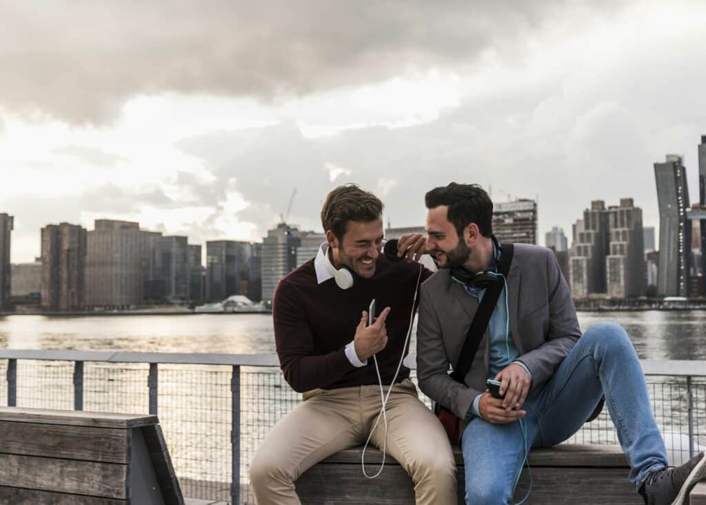 USA, New York City, two happy young men with headphones and cell phone sitting at East River